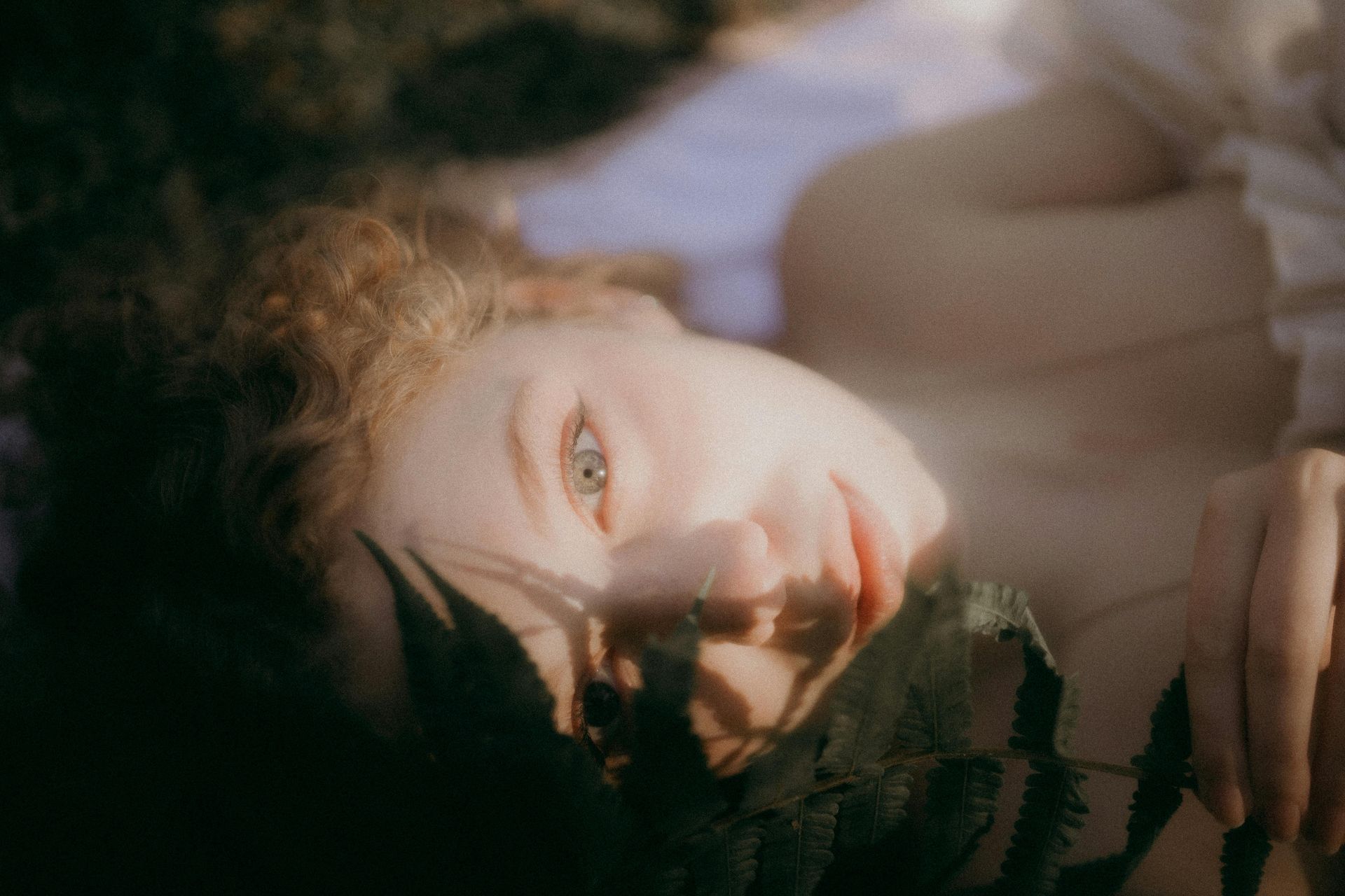 Woman lying in sunlight, eyes open. Fern fronds partially obscure face, soft focus, warm tones.