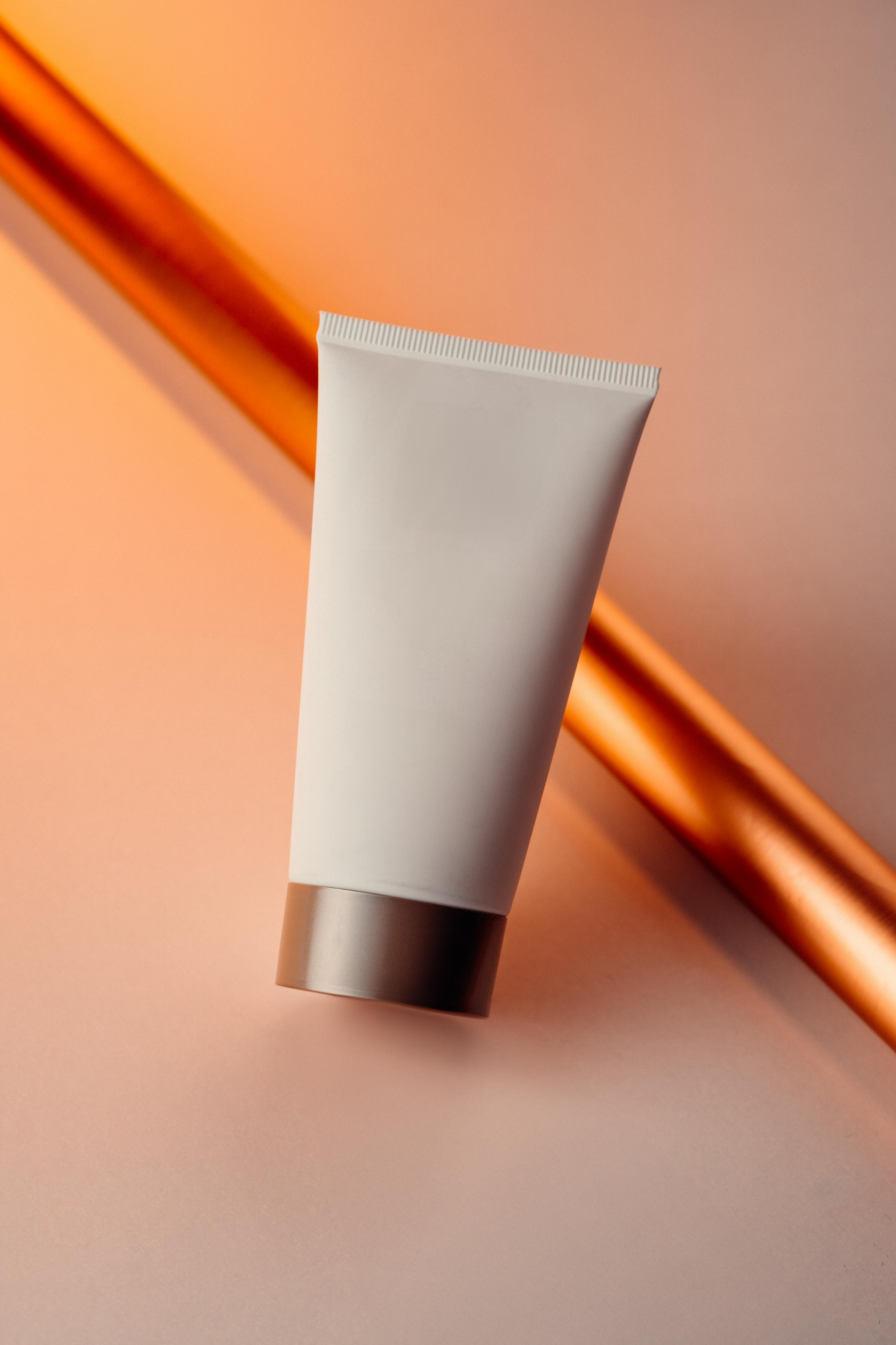 White cosmetic tube, brown cap, angled against a copper-colored rod on a peach background, lit by warm light.