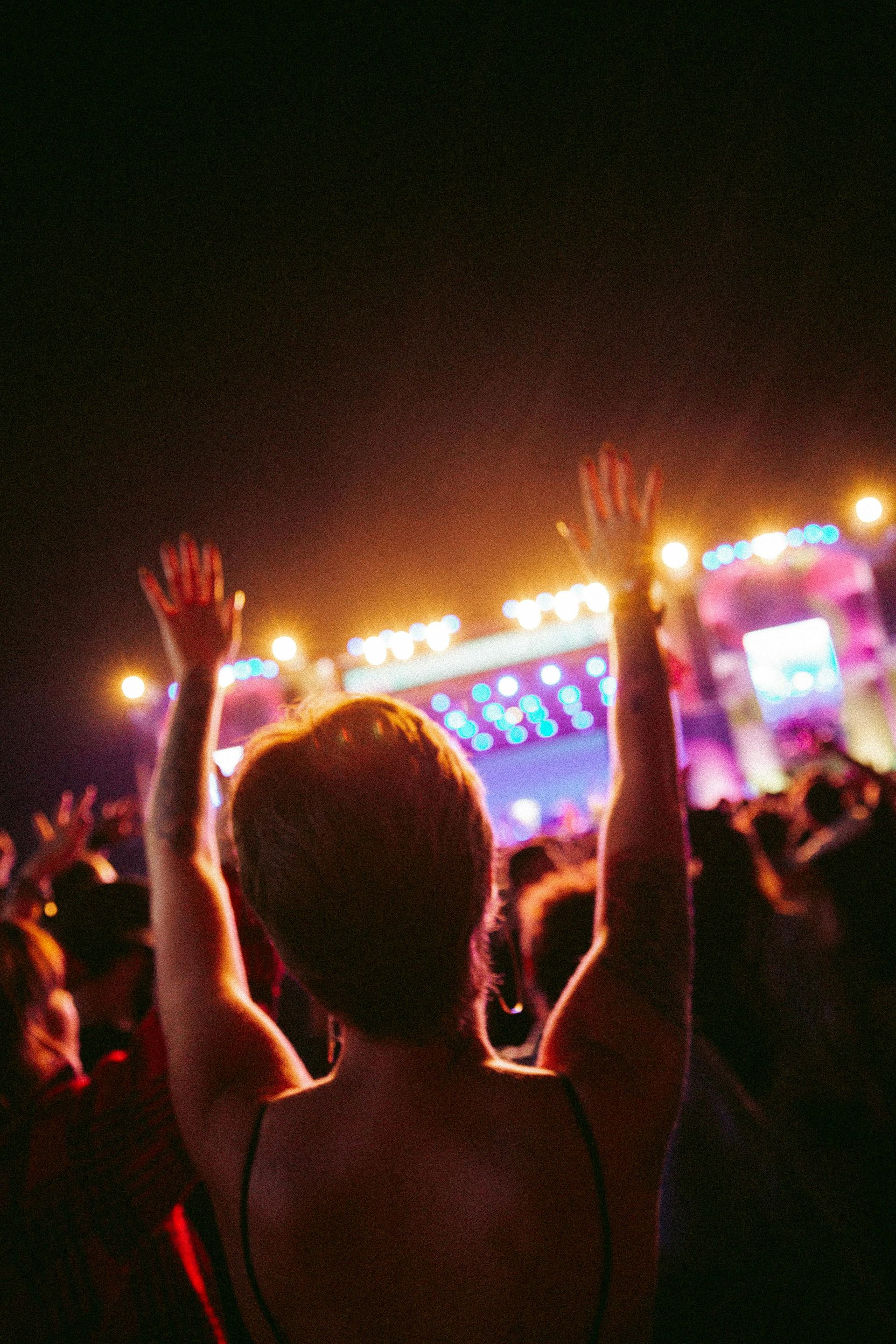 Person with arms raised at concert, stage with lights in background.