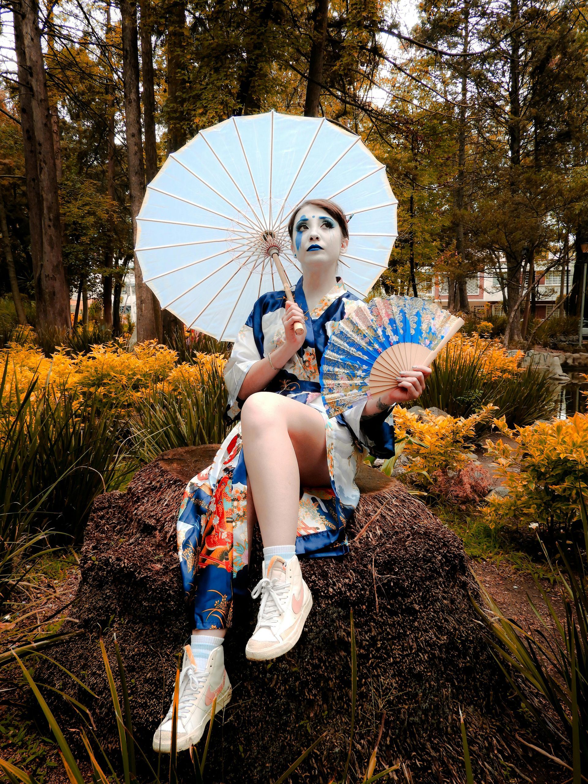 Person in blue kimono, holding parasol and fan, seated on rock in a park setting.