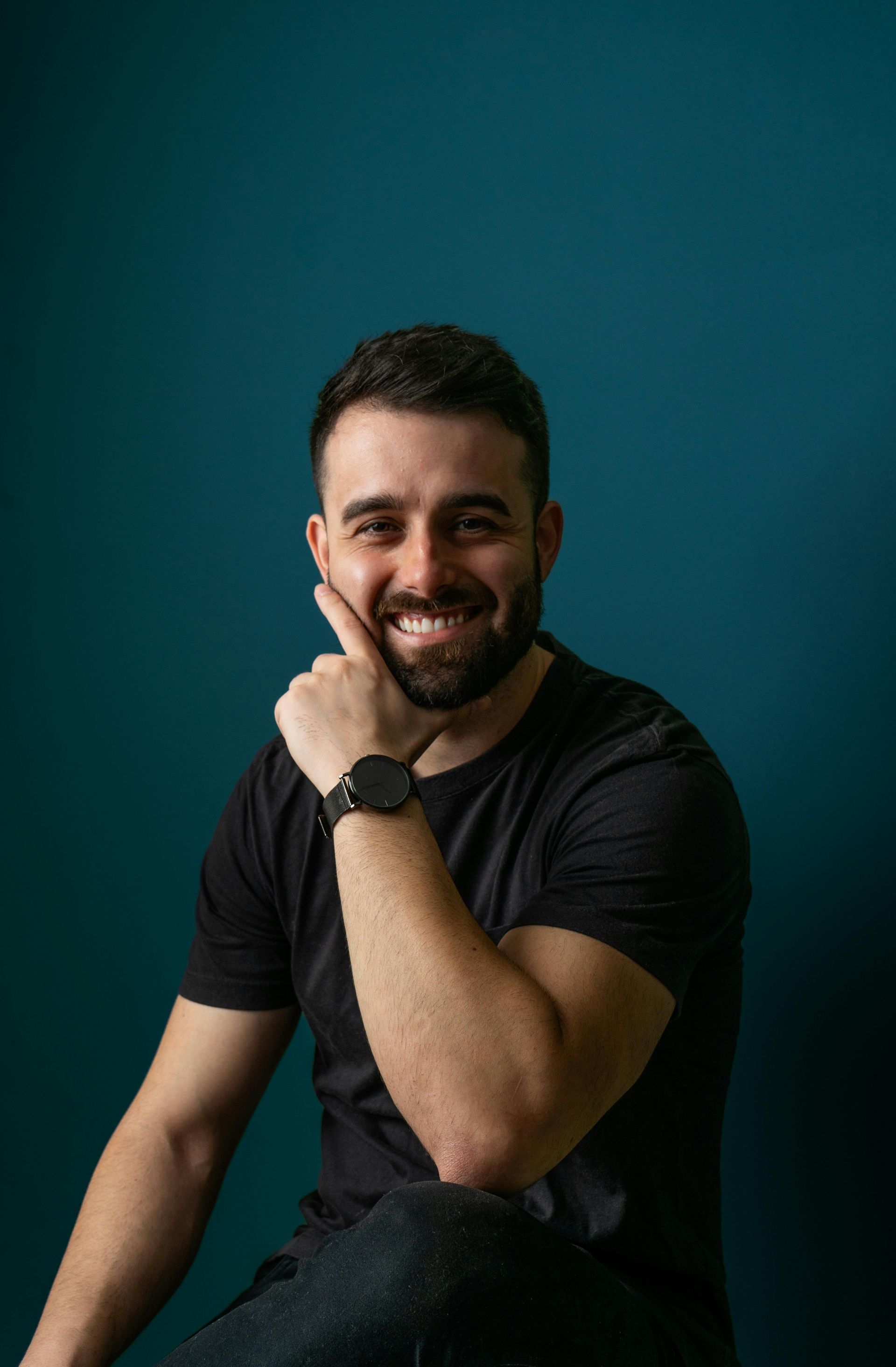 Man with beard, smiling, resting hand on cheek. Wearing black shirt, watch, sitting against teal backdrop.