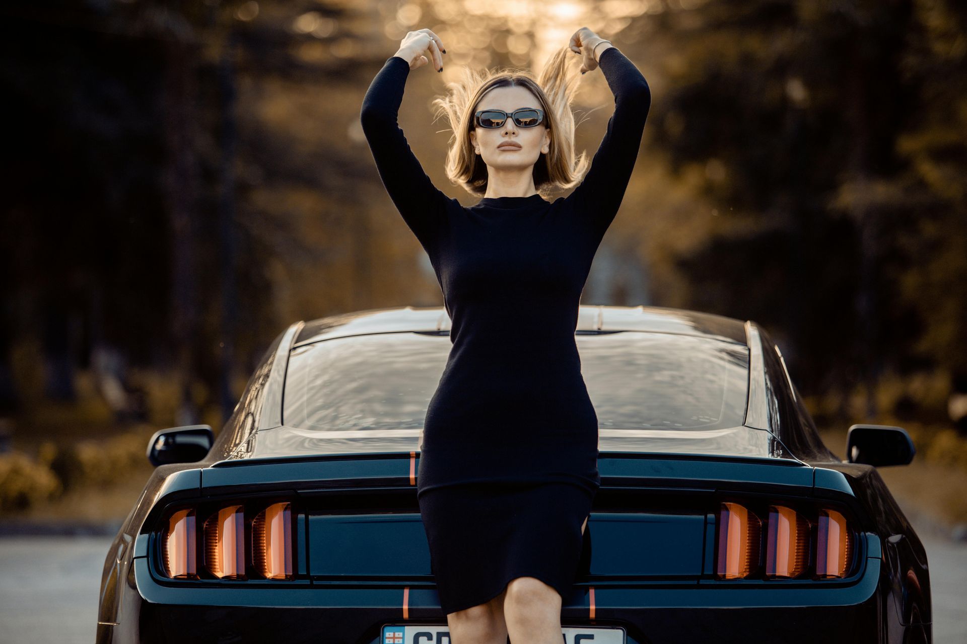 Woman in black dress and sunglasses poses with arms raised in front of a black sports car.