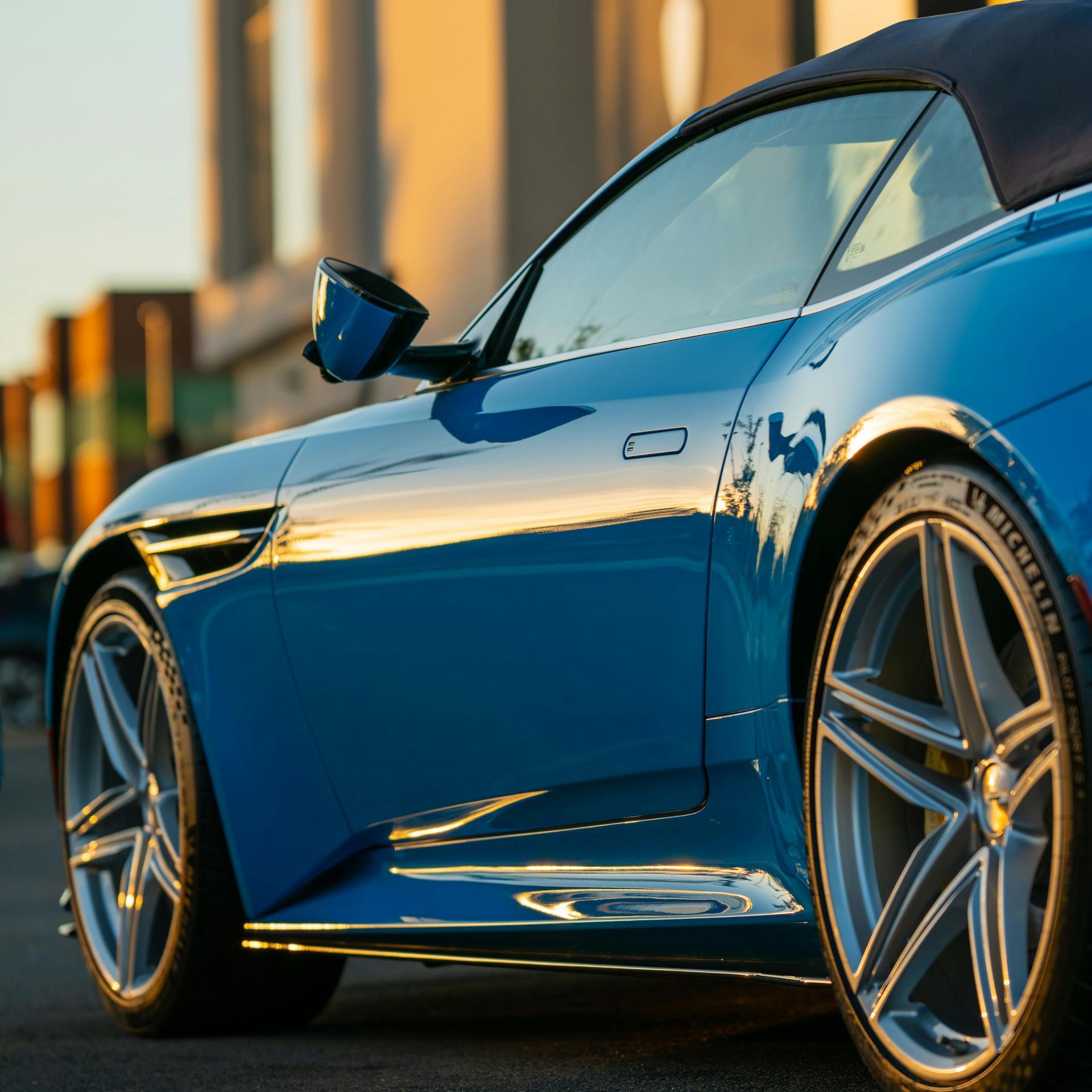 Blue convertible sports car with silver wheels parked on a street; reflected sunlight.