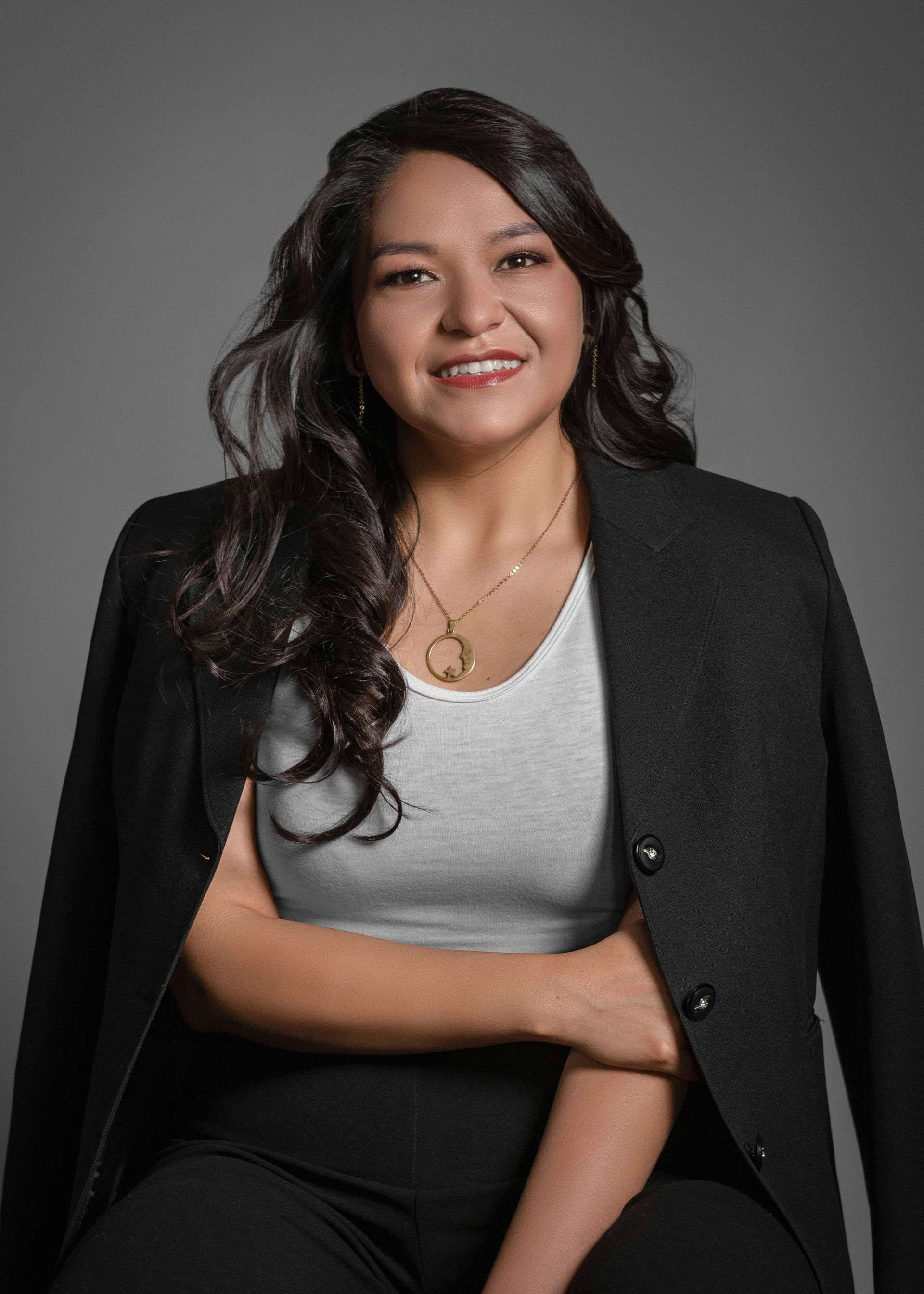 Woman in black blazer over a white top, smiling. Sitting against a gray backdrop.
