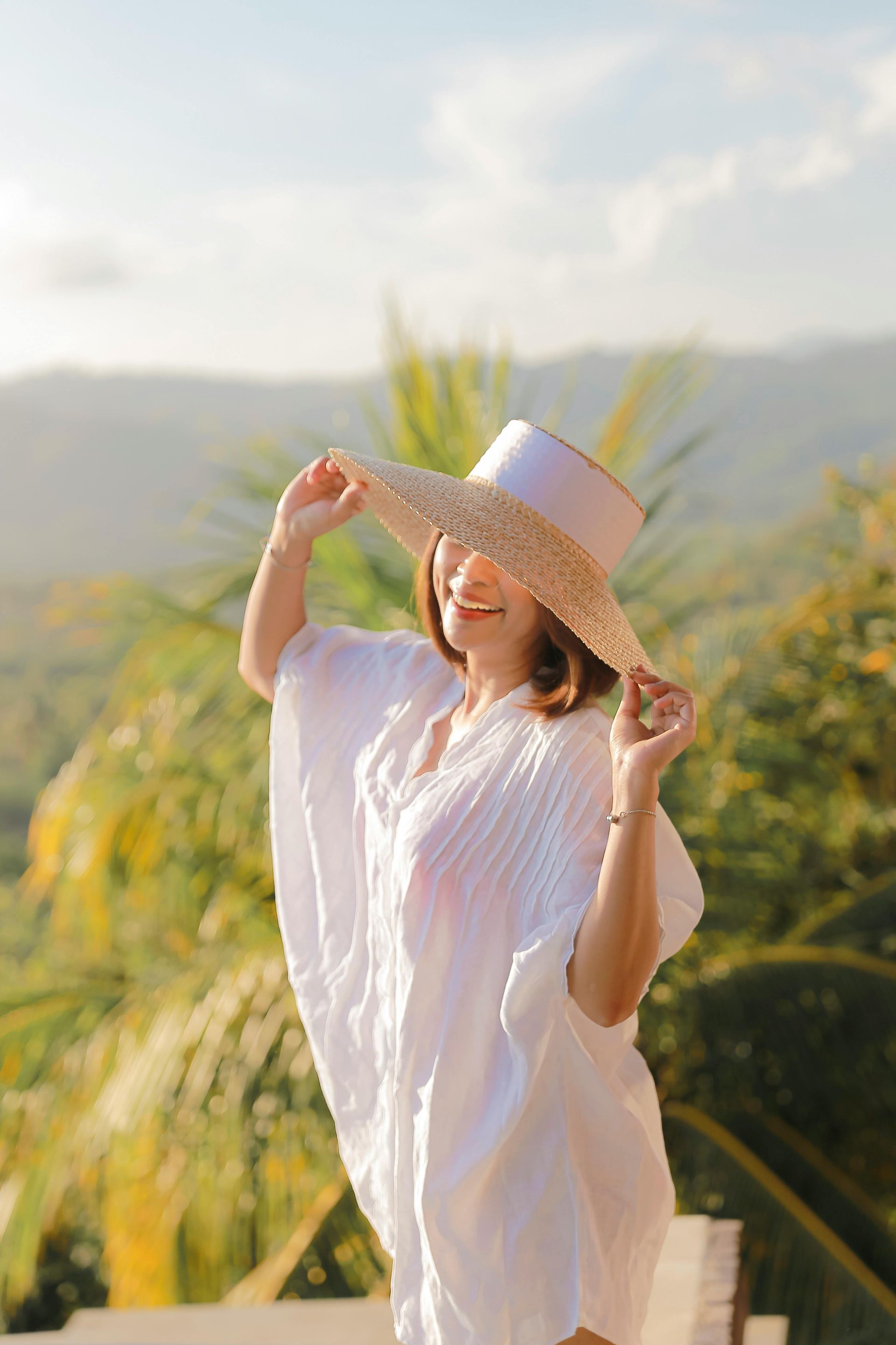 Woman smiles, holding a sun hat, wearing a white cover-up outdoors with green foliage and mountains in the background.