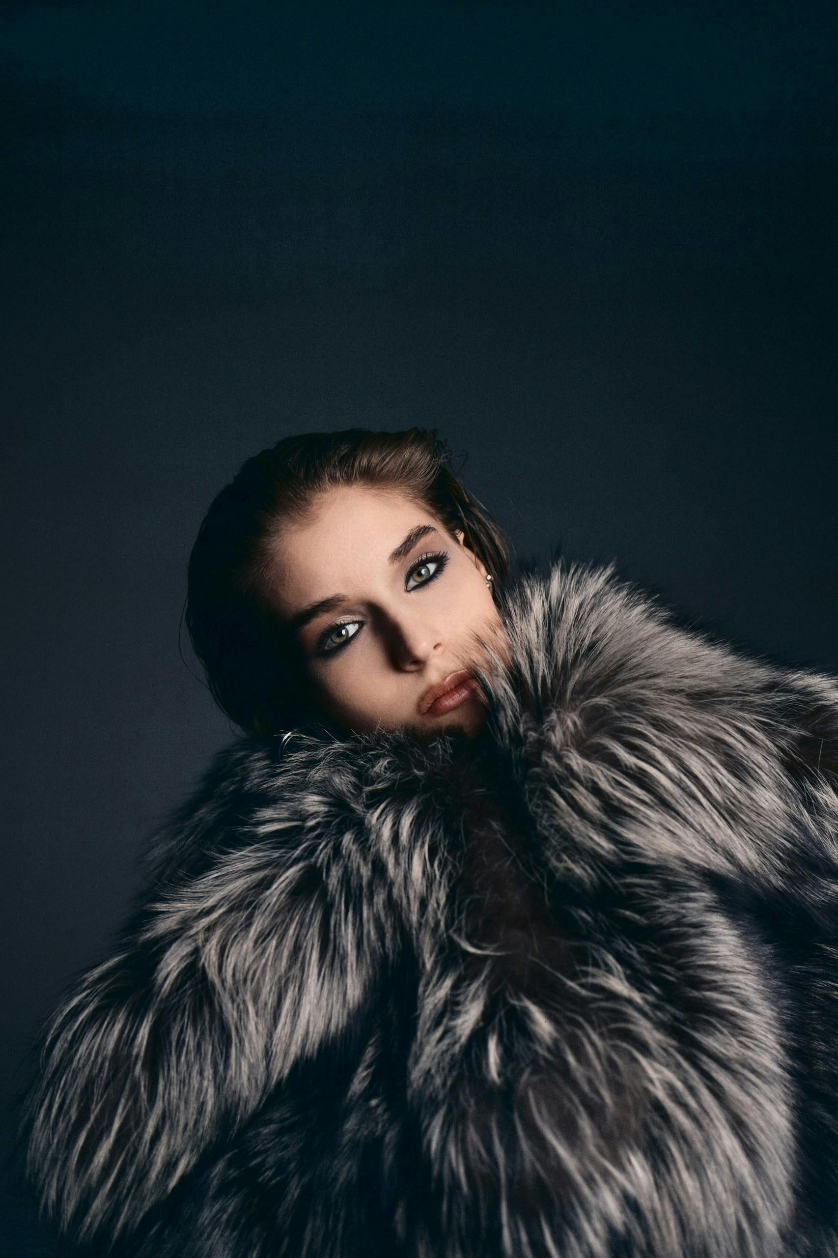 Woman in silver fur coat, looking towards the viewer with dark eye makeup and red lipstick, against a dark background.