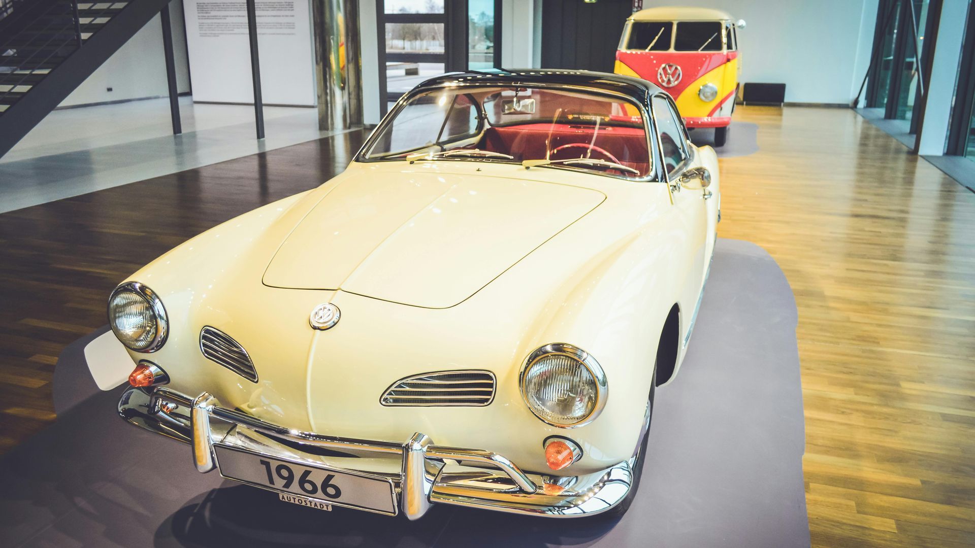 Cream vintage convertible on display indoors, with a yellow-and-red VW van in the background.