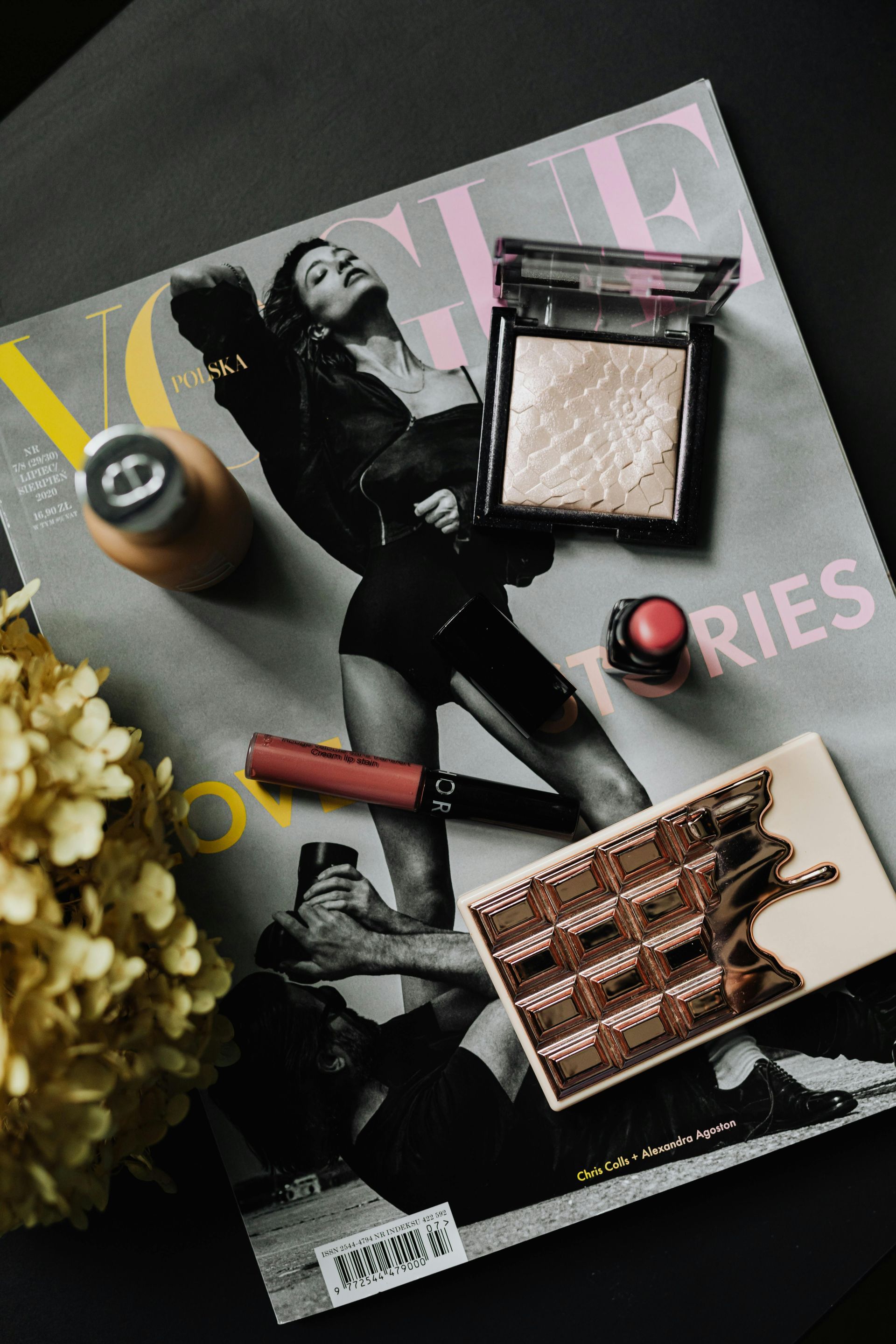 Makeup products arranged on an open Vogue magazine; cosmetics include highlighter, lipstick, and eyeshadow palette.