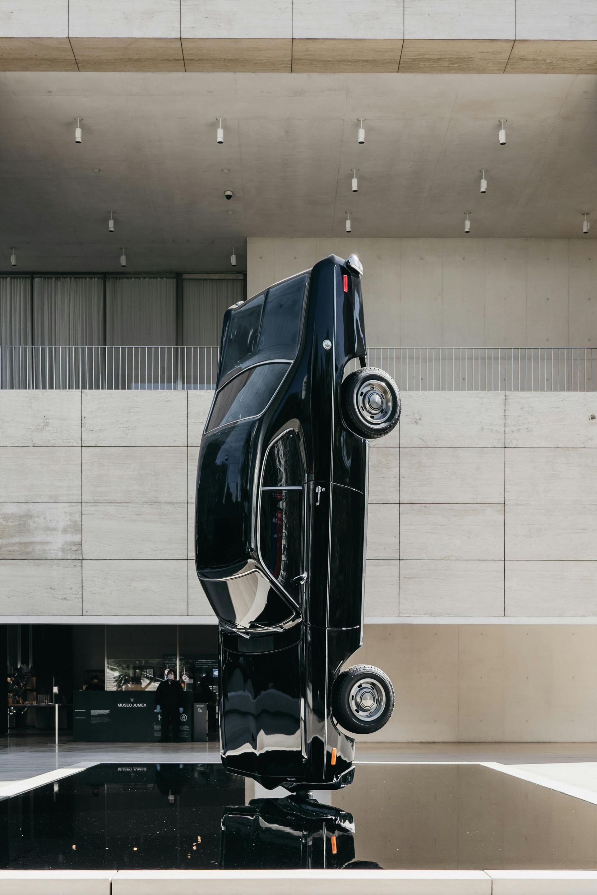 Black upside-down car sculpture reflected in dark water, outside a modern building with light walls.