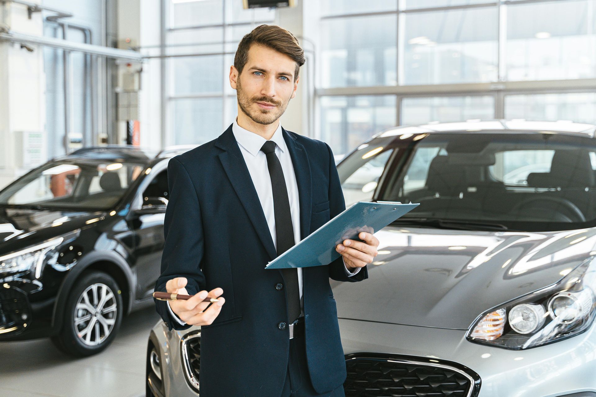 Man in a suit holding a clipboard and pen stands in a car dealership, cars in background.