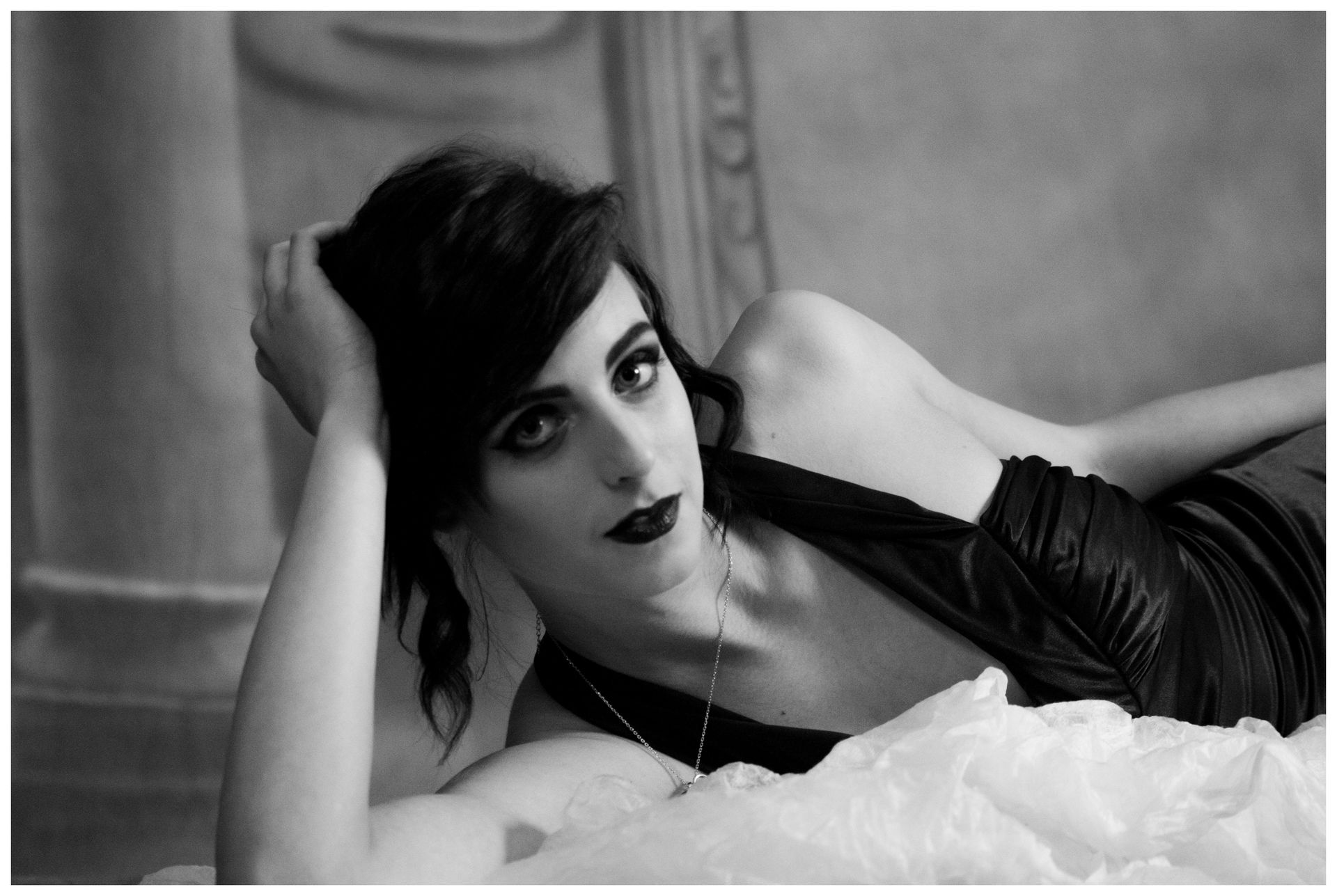 Woman in a dark dress reclines on a bed, looking towards the viewer with dark lipstick.