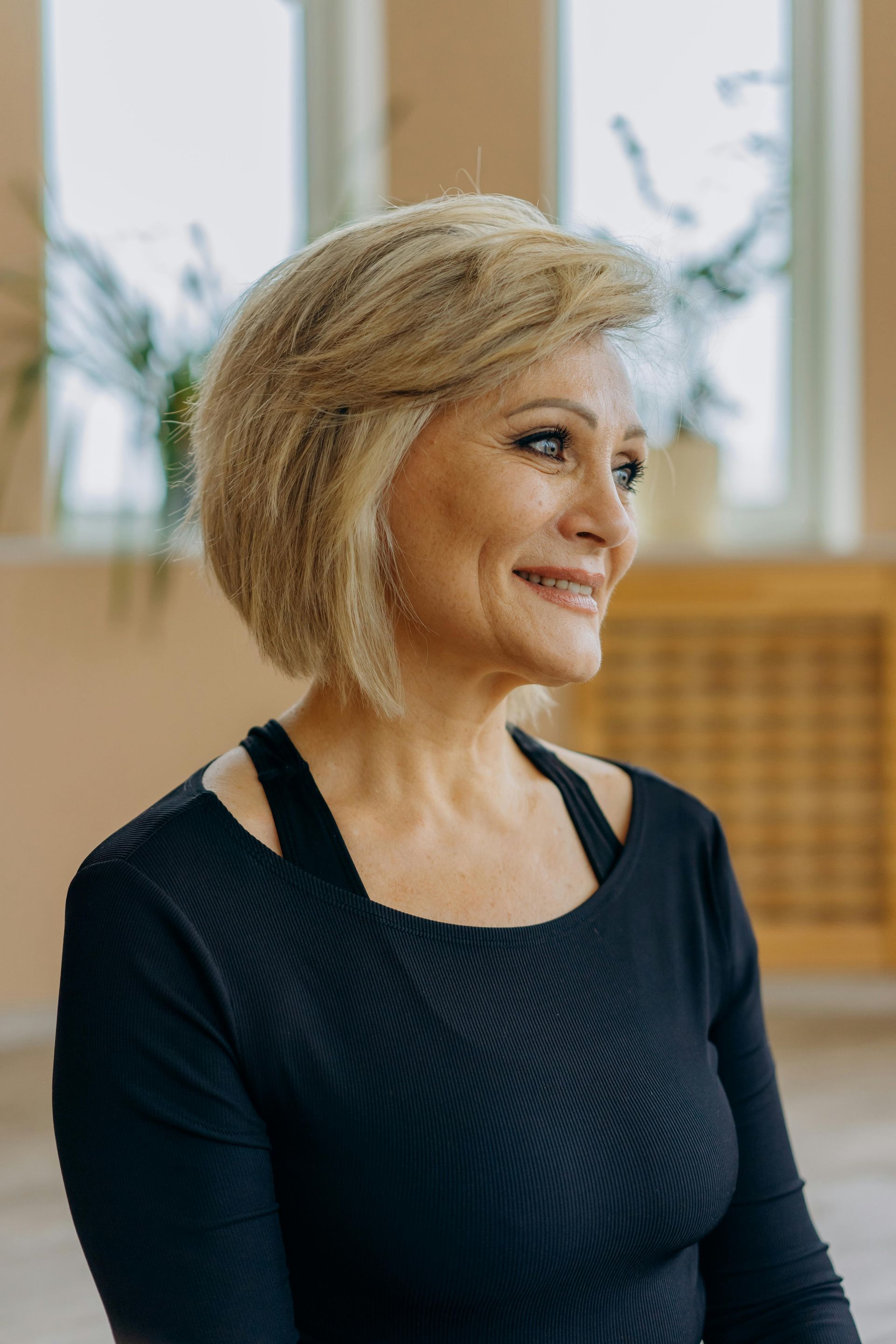 Woman with short, light-colored hair, wearing a black top, smiling in a room with windows.