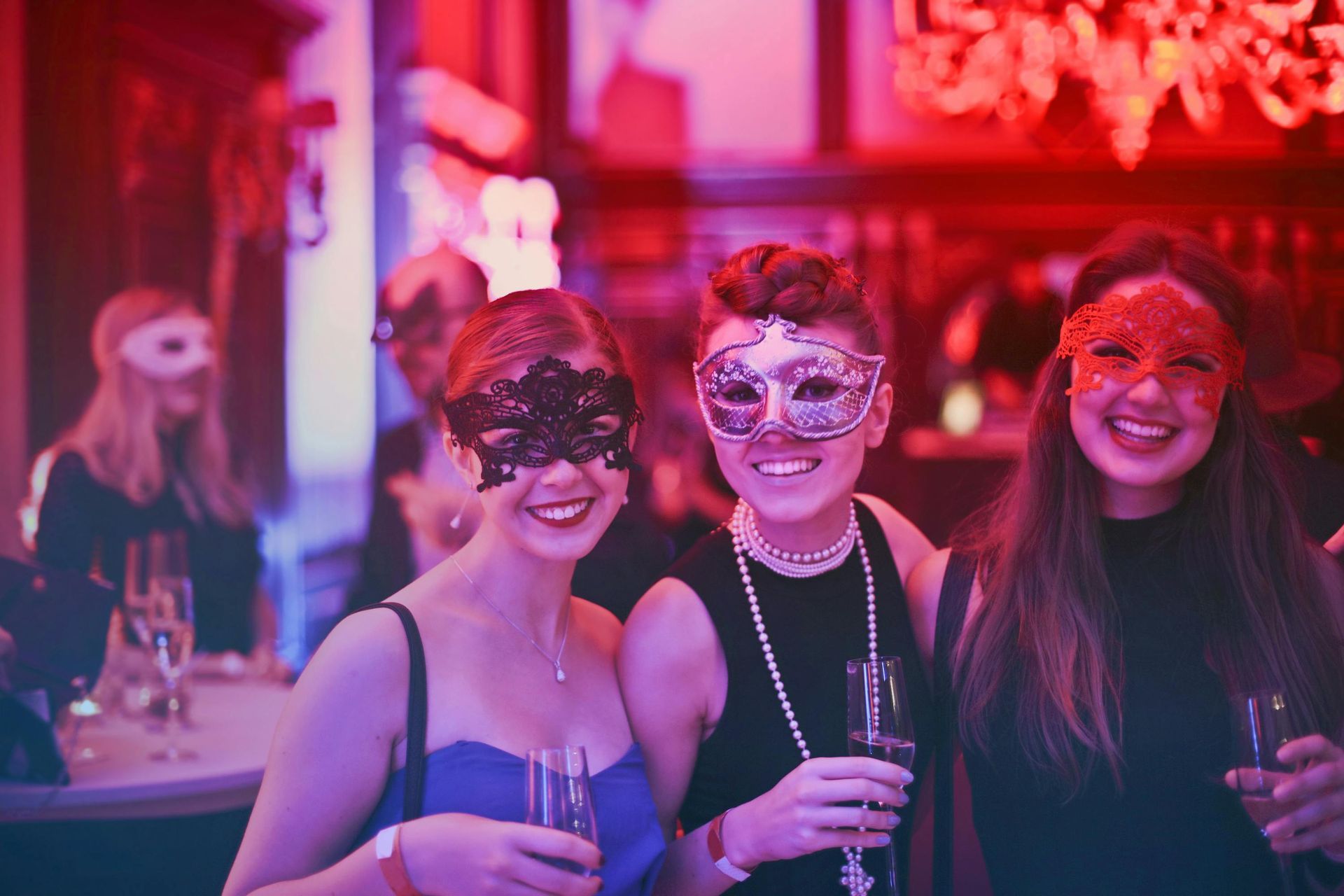 Three people wearing masks at a party, holding glasses, smiling. Red and blue lighting.