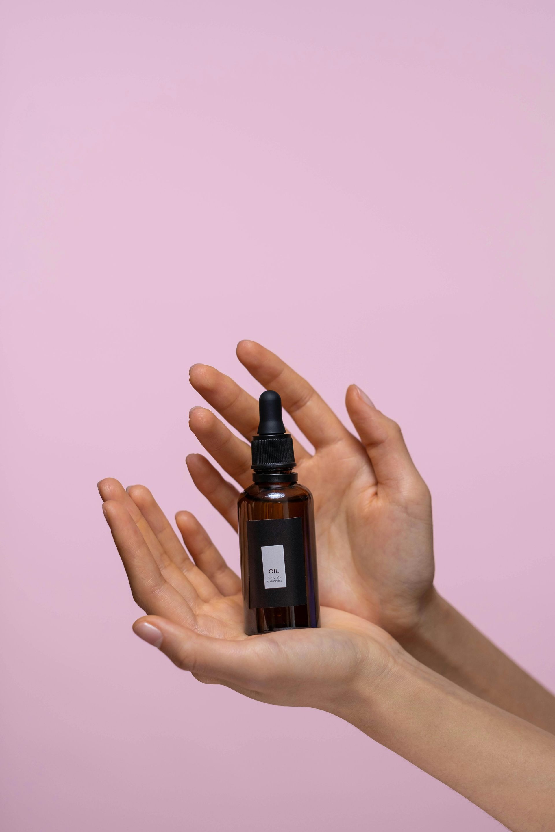 Hands holding a brown dropper bottle with a black label, on a pink background.