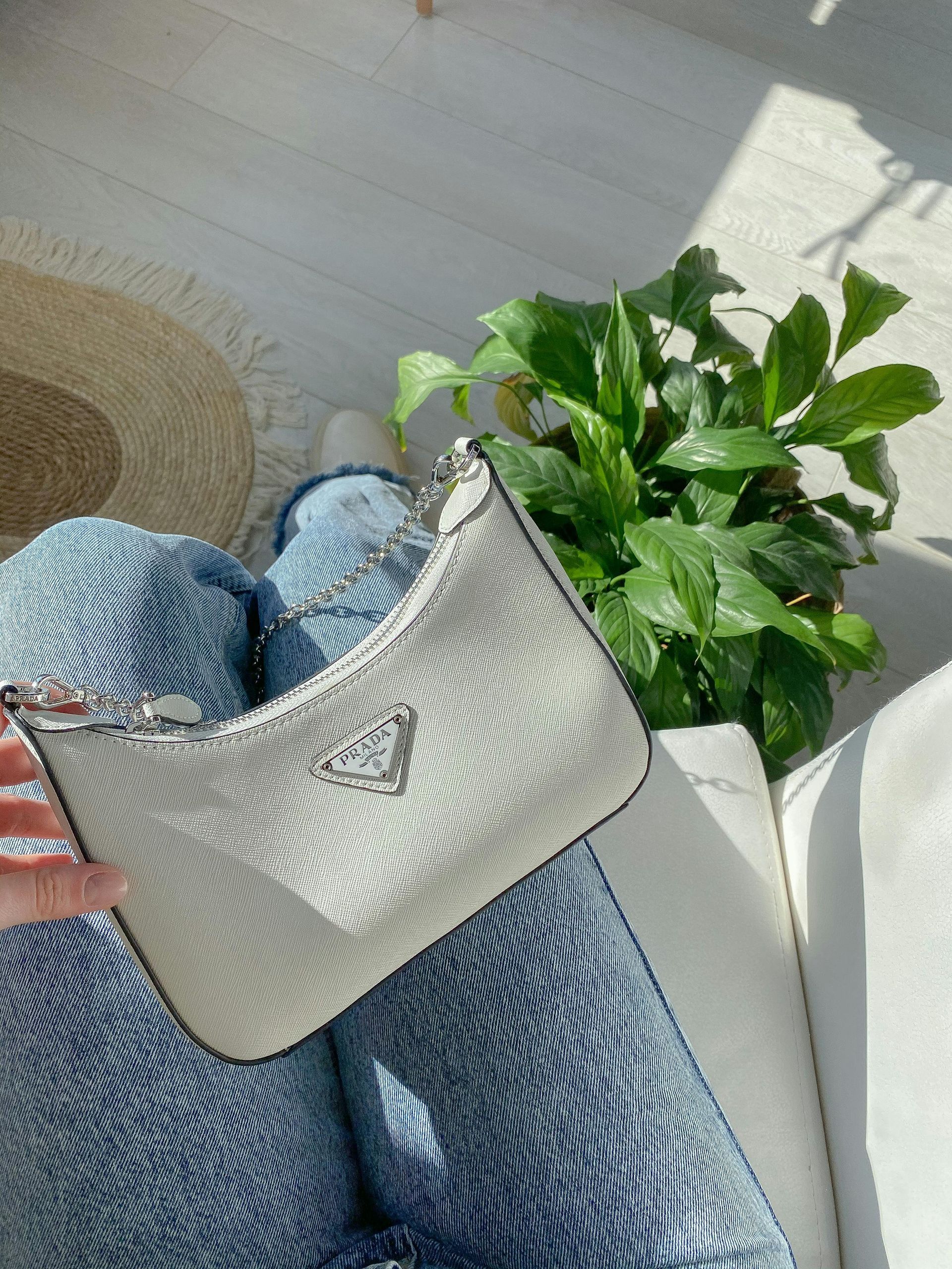 Silver Prada handbag on denim-clad legs near houseplant. Bright sunlight.