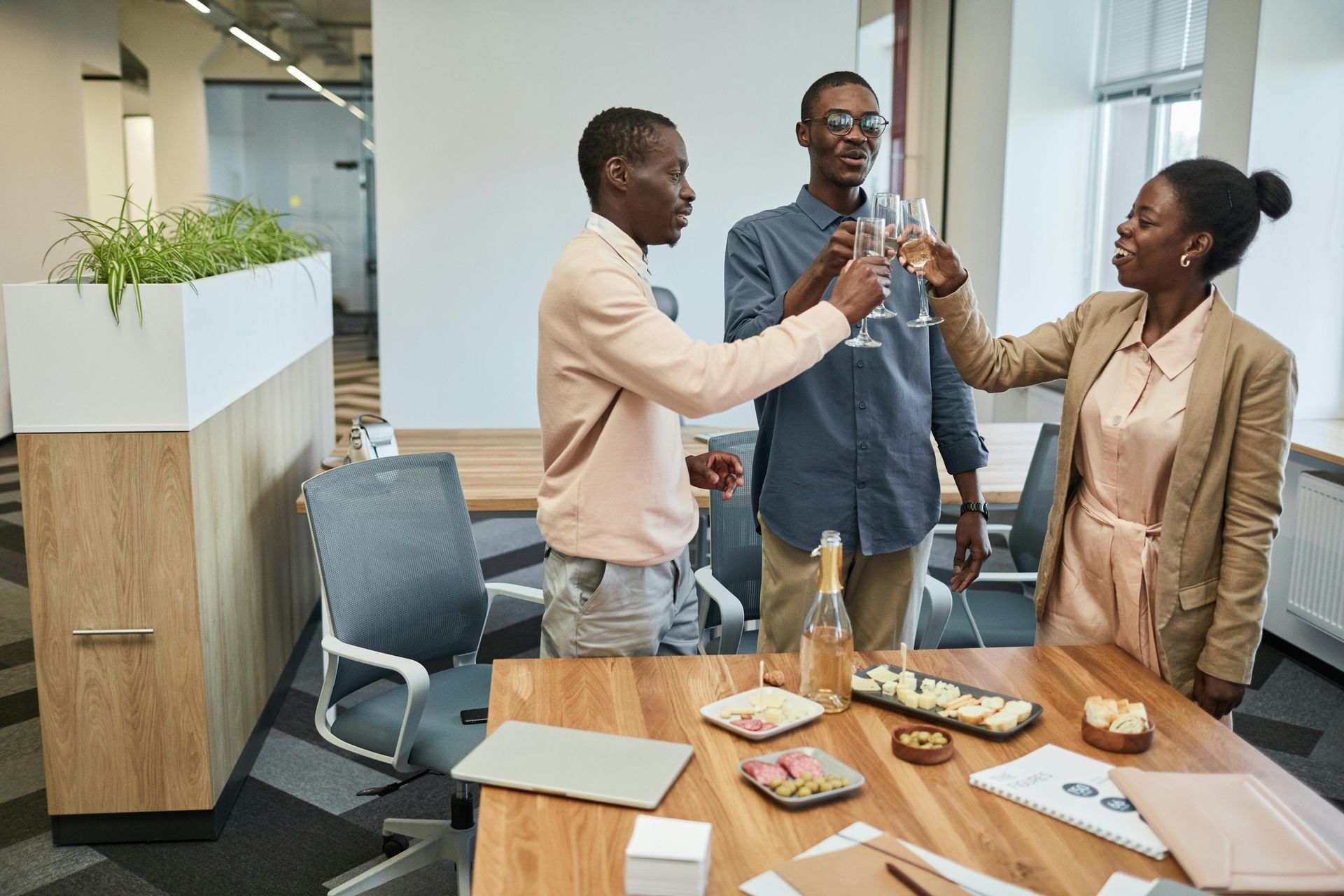 Three people in an office toasting with glasses. Snacks and papers on a table.