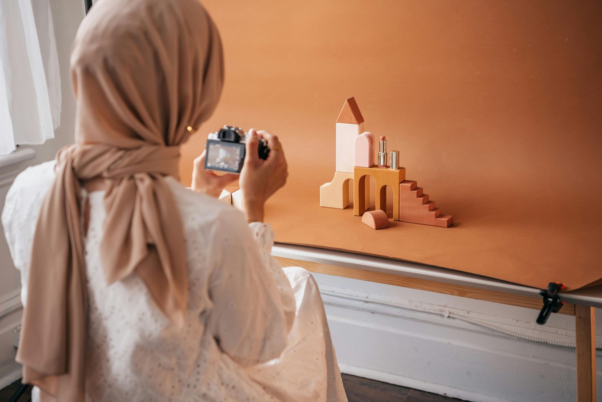 Woman in hijab photographing product arrangement on a table with a brown backdrop.