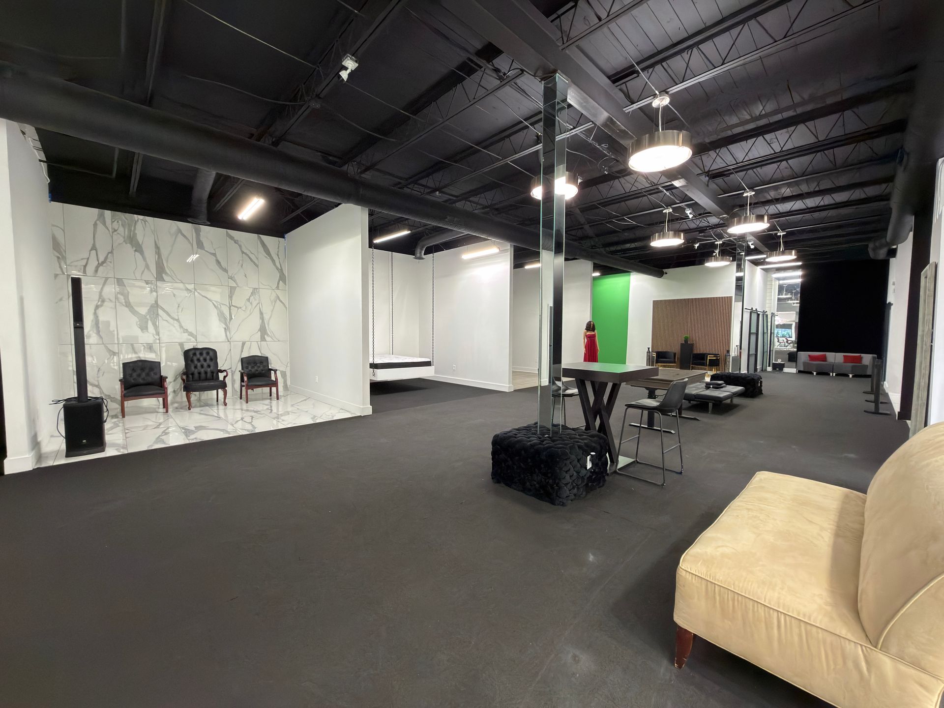 Spacious empty studio with black floor, white walls, exposed ceiling, chairs, tables, and a tan couch.