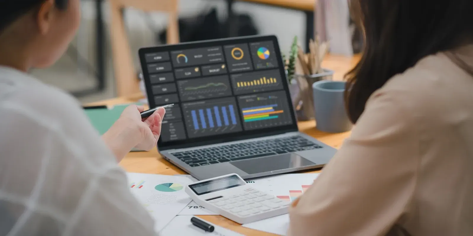 Two women analyzing data on a laptop. Charts, calculator, and papers on a desk.