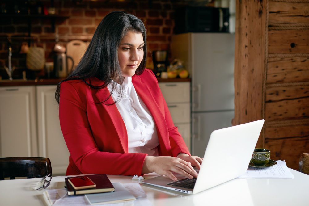 A woman in a red jacket is sitting at a table using a laptop computer.