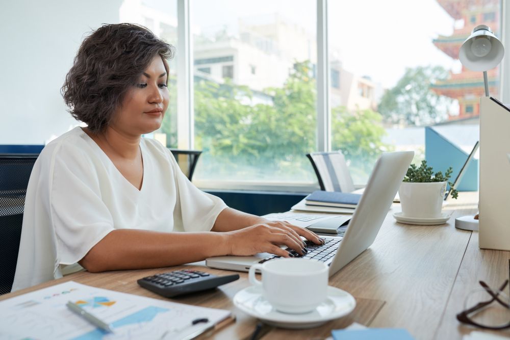 A woman is sitting at a desk typing on a laptop computer.