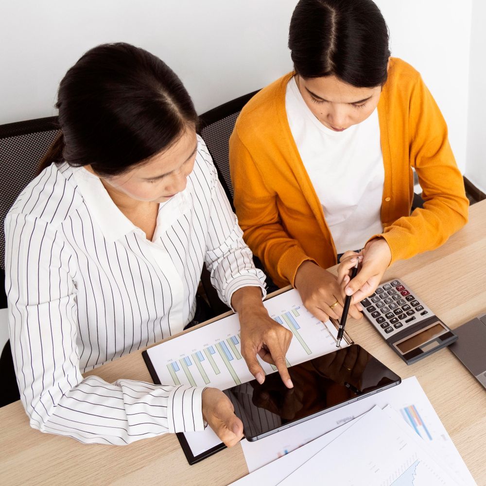 Two women are sitting at a table looking at a tablet and a calculator.