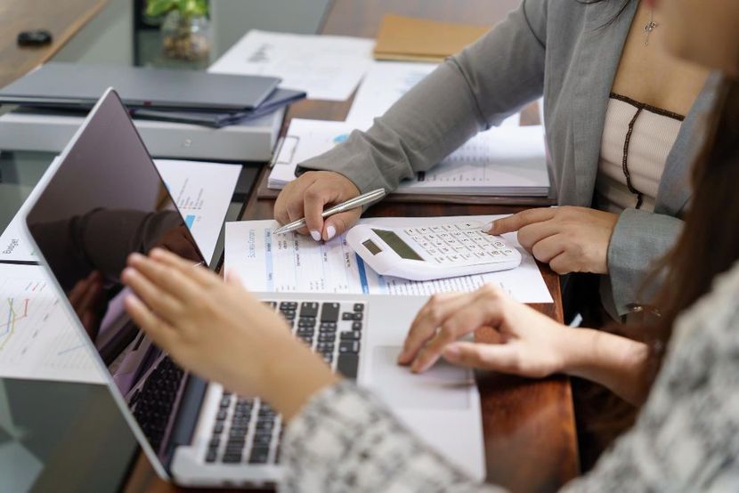 Two women are sitting at a table using laptops and a calculator.