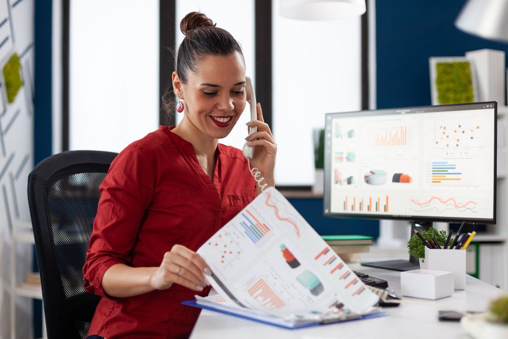 A woman is sitting at a desk talking on a phone while holding a clipboard.