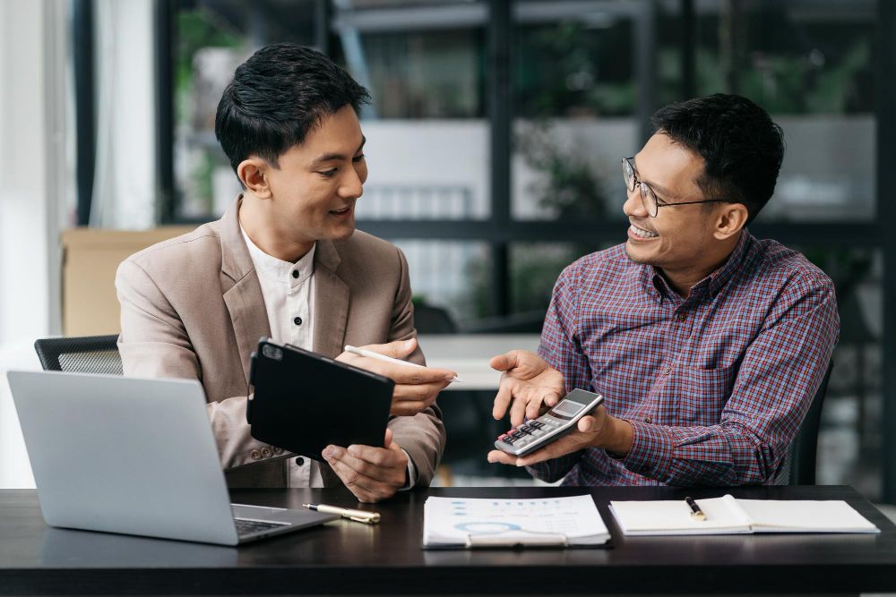 Two men are sitting at a table with a laptop and a calculator.
