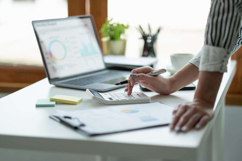 A woman is using a calculator at a desk in front of a laptop.