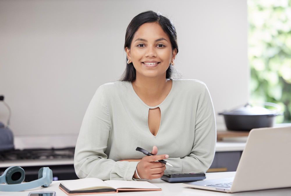 A woman is sitting at a desk with a laptop and a notebook.