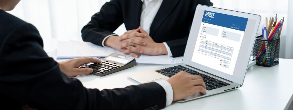 A man is typing on a laptop while a woman is sitting at a desk with a calculator.