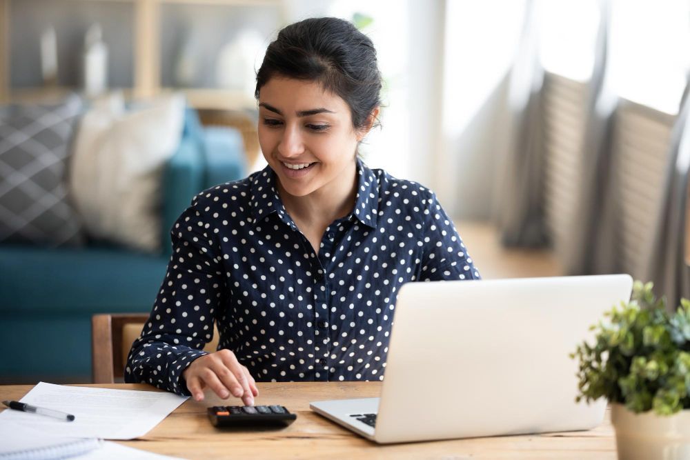 A woman is sitting at a table using a laptop computer and a calculator.