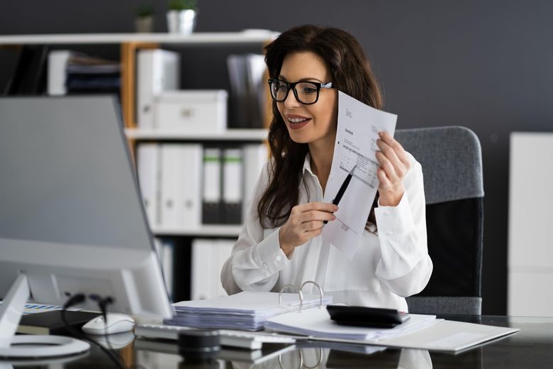 A woman is sitting at a desk in front of a computer holding a piece of paper.