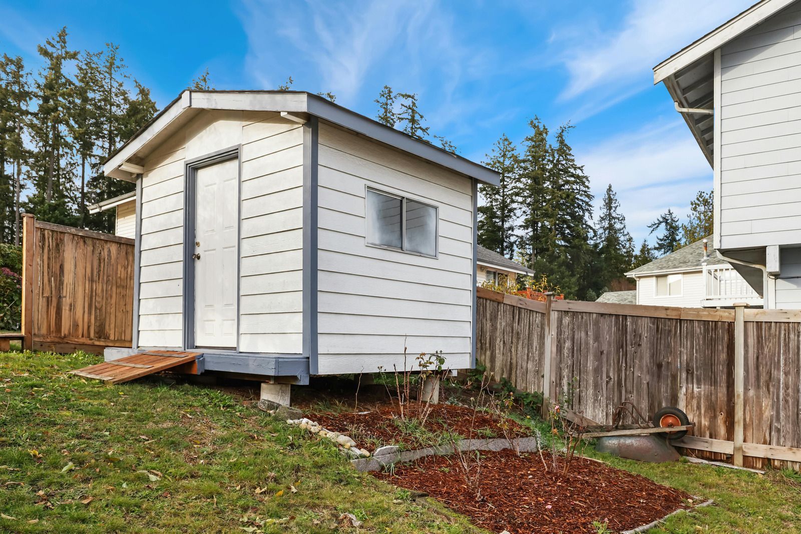 Small white shed with blue trim, wooden fence, and landscaping in a yard.