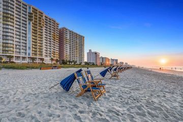 Beach chairs on a sandy shoreline beside high-rise condos at sunset, with the ocean and warm sky in view.
