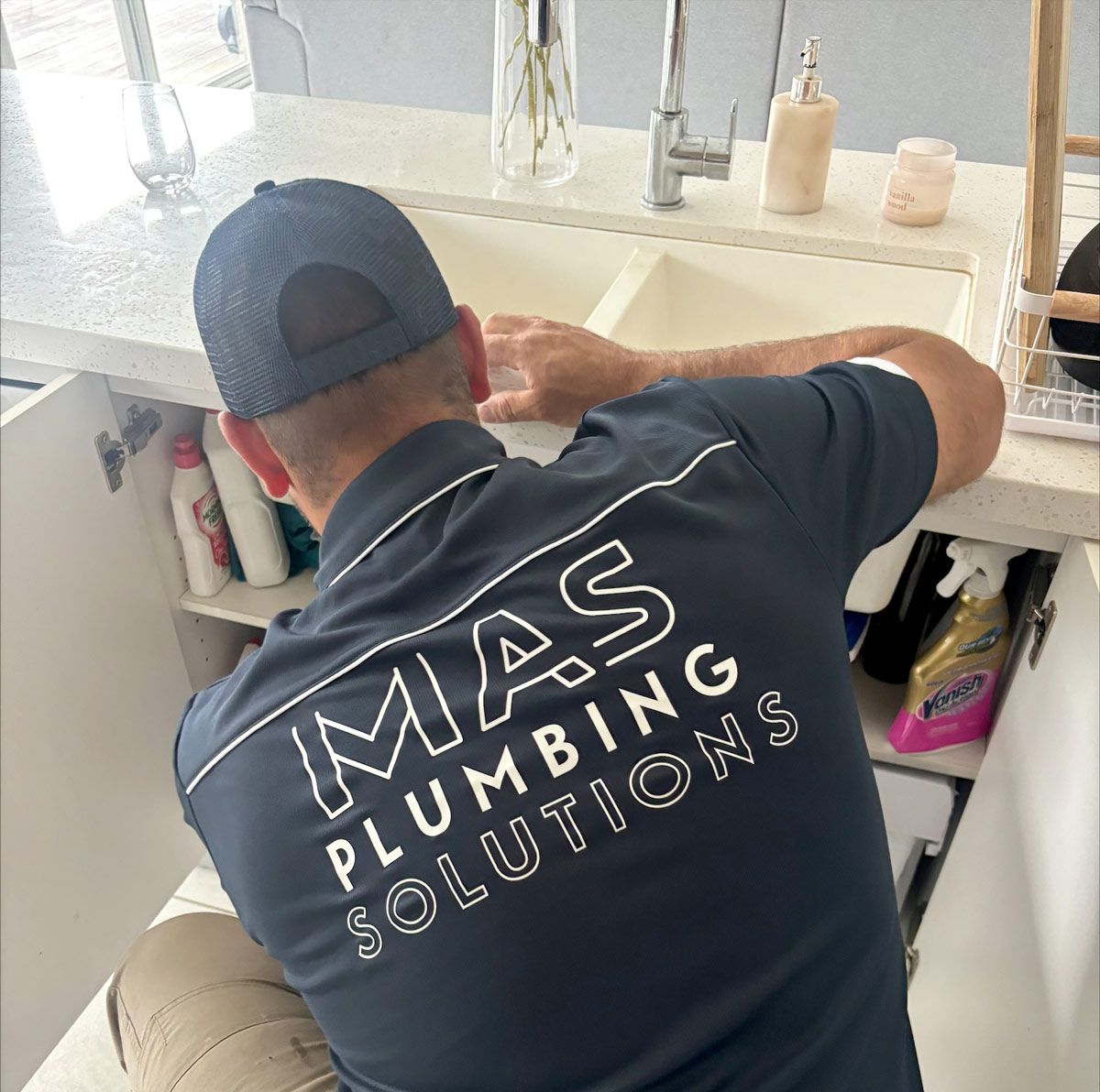 A man wearing a mas plumbing solutions shirt is working on a sink