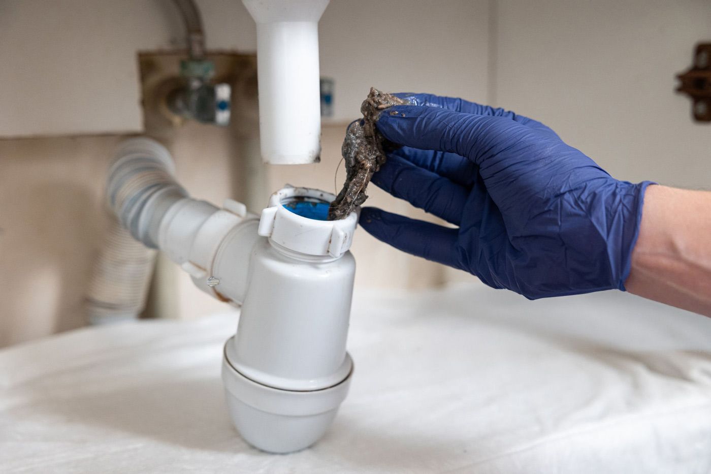 A gloved hand pulls a dark, matted clog of debris out of a disassembled white plastic sink drain trap.