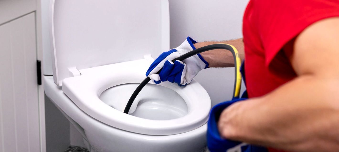 A person in a red shirt and work gloves uses a plumbing snake to clear a clog in a white toilet bowl.