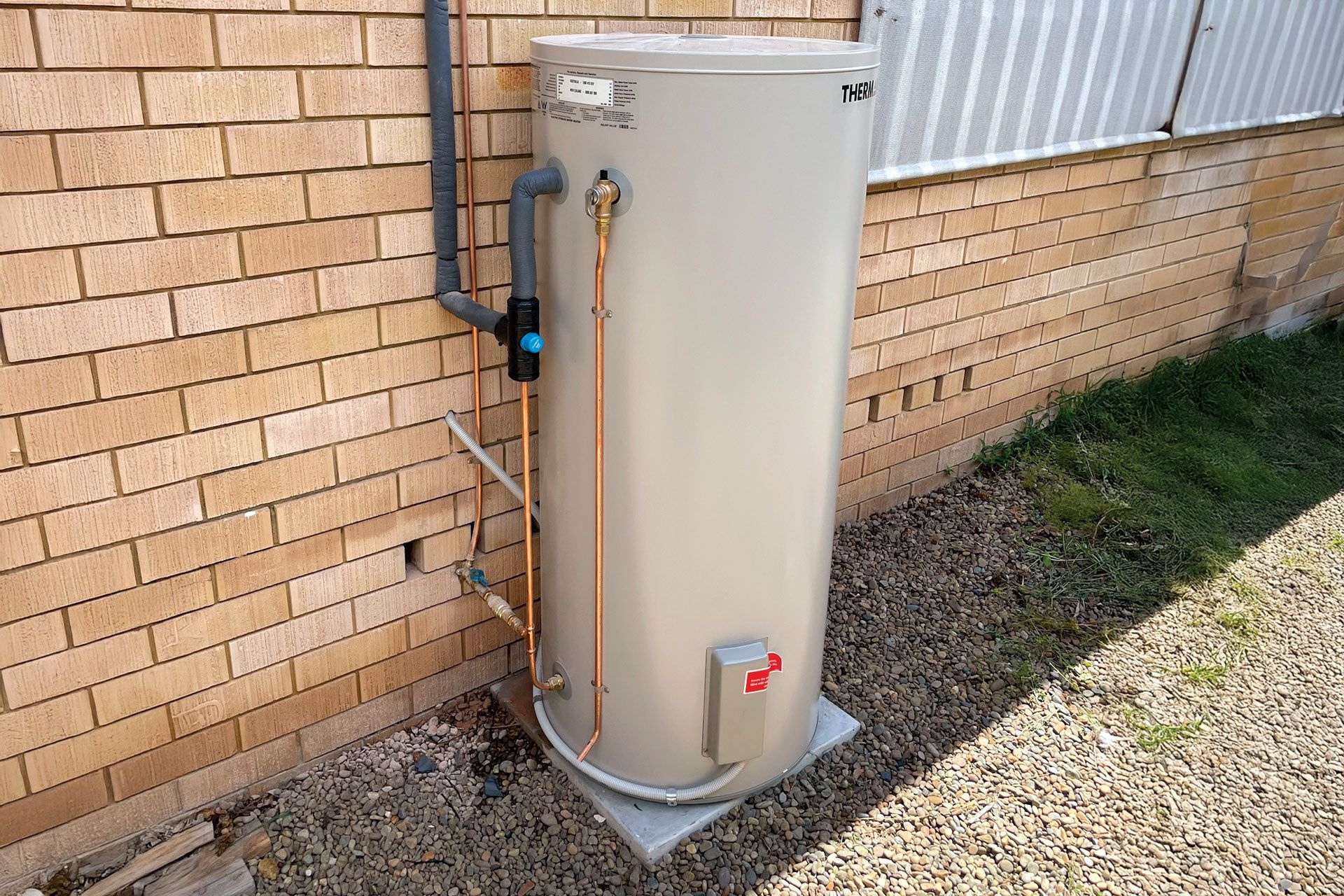 A beige cylindrical water heater stands against a brick wall on a gravel surface with attached copper plumbing pipes.