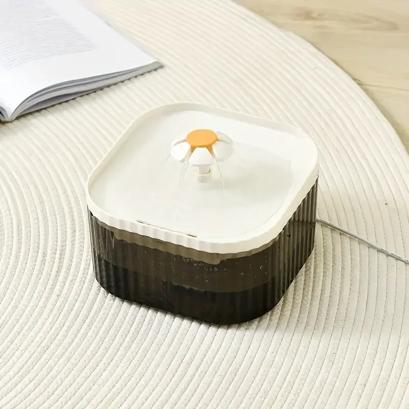 Fuente de agua para mascotas con caño en forma de margarita, en blanco y negro, colocada sobre una alfombra blanca con un libro.