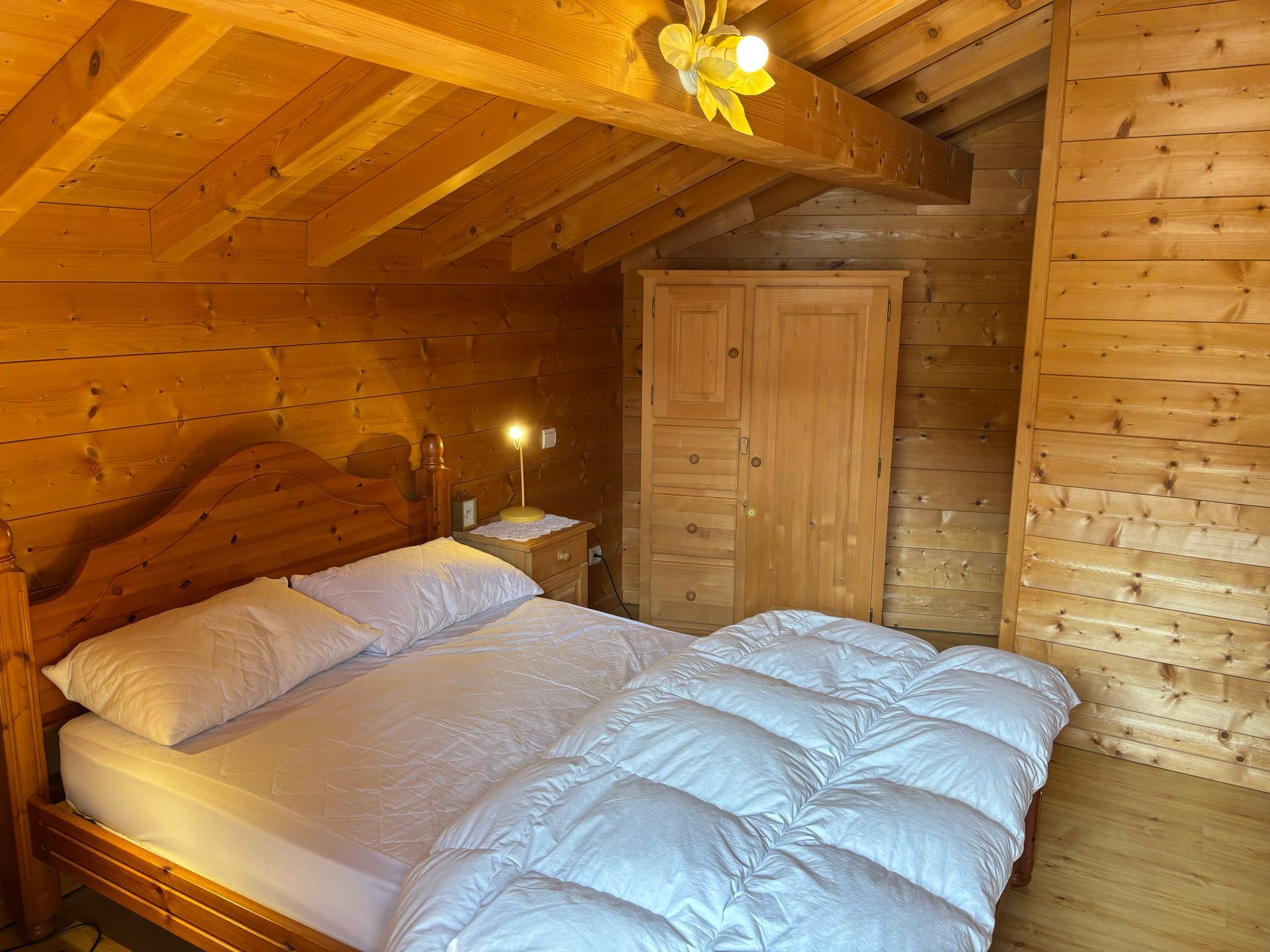 Bedroom with wood-paneled walls, ceiling, and bed frame. A bed has a white duvet and pillows. A wooden wardrobe and nightstand with lamp are visible.
