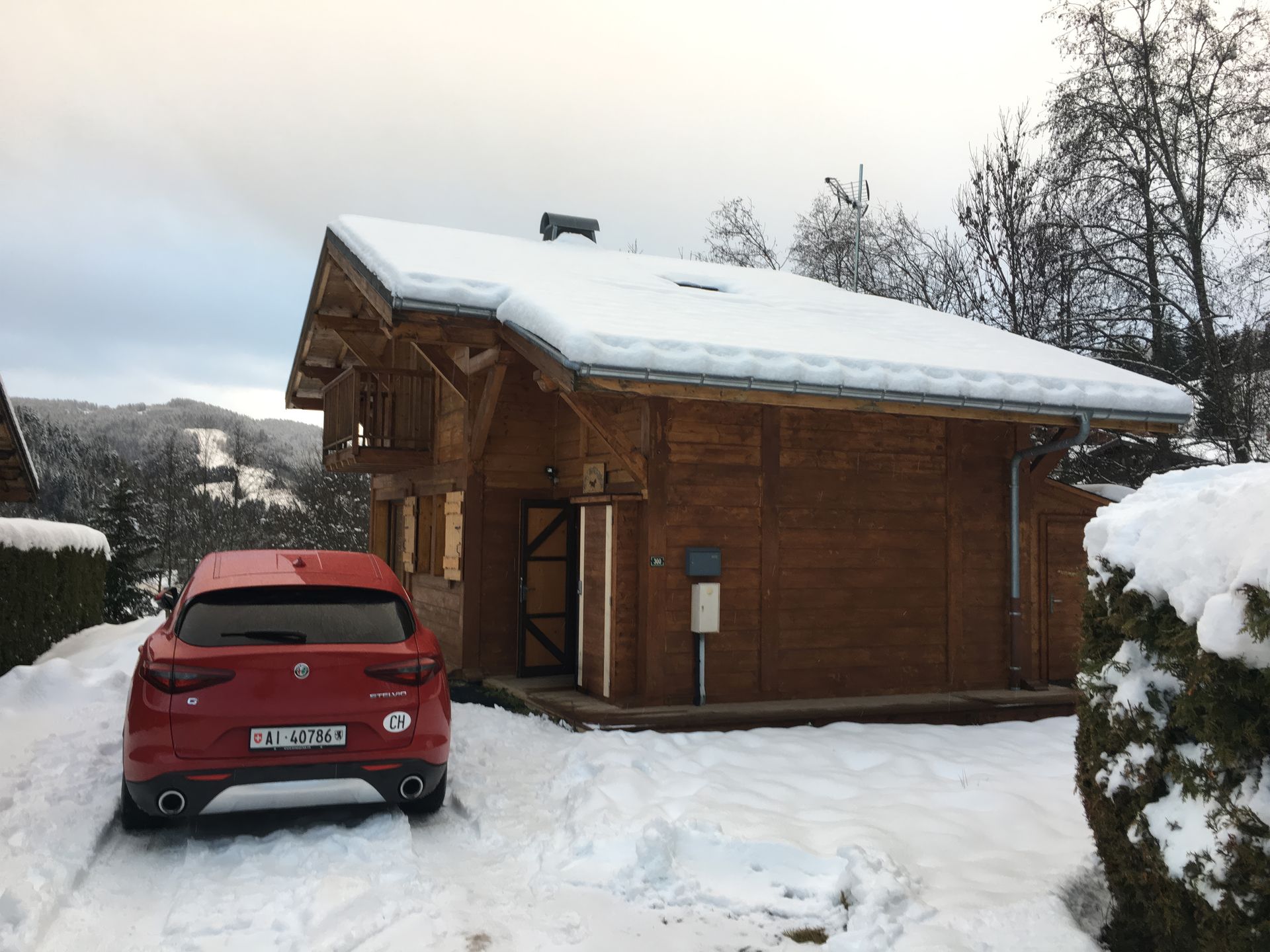 Red car parked outside a wooden cabin covered in snow. Mountain view in background, cloudy sky.