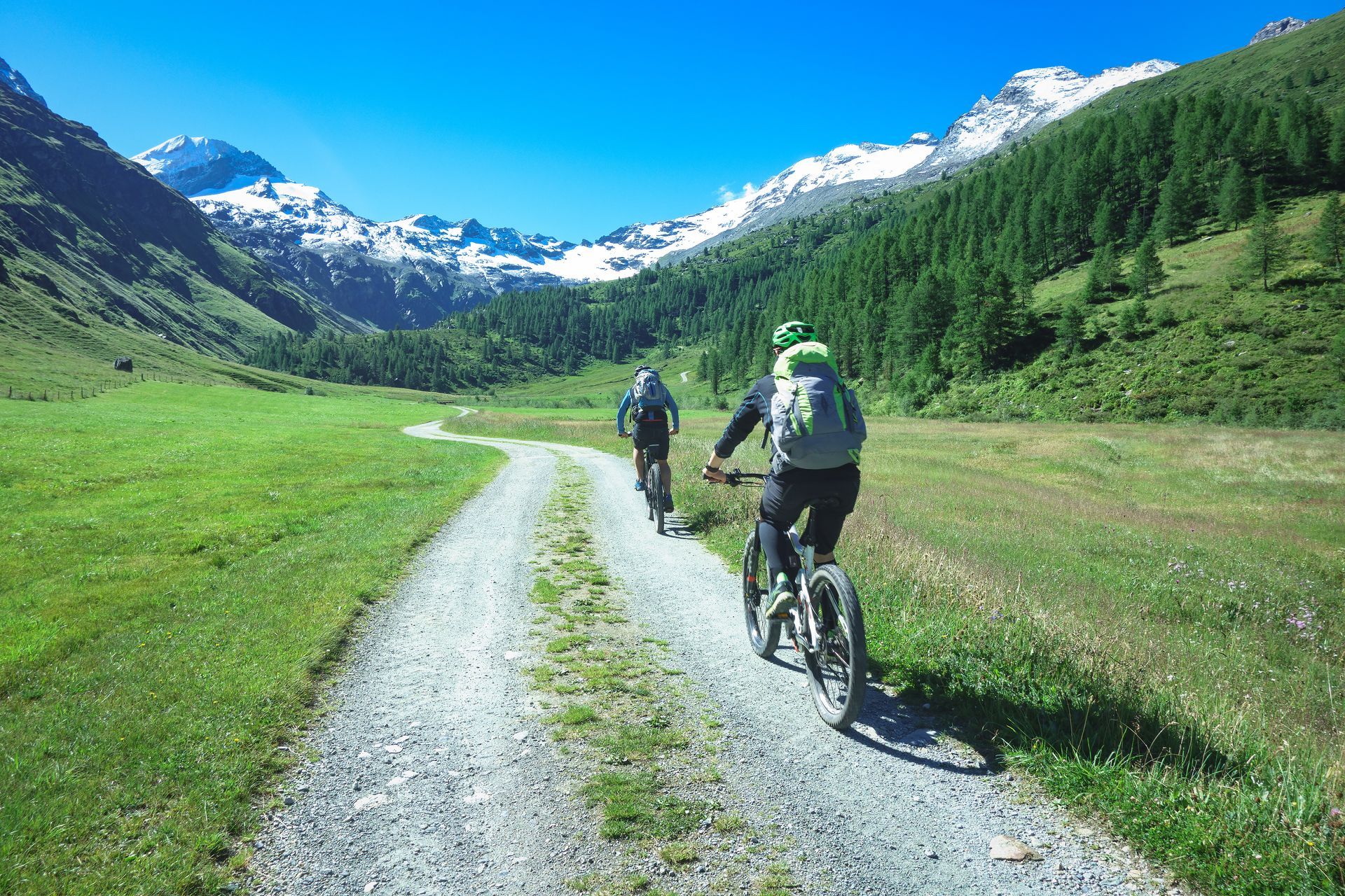 Two cyclists ride on a gravel path in a green valley, mountains in the background under a blue sky.