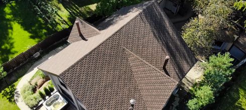 Vue aérienne d'une maison avec un toit à motifs bruns entouré d'arbres verts et d'herbe.