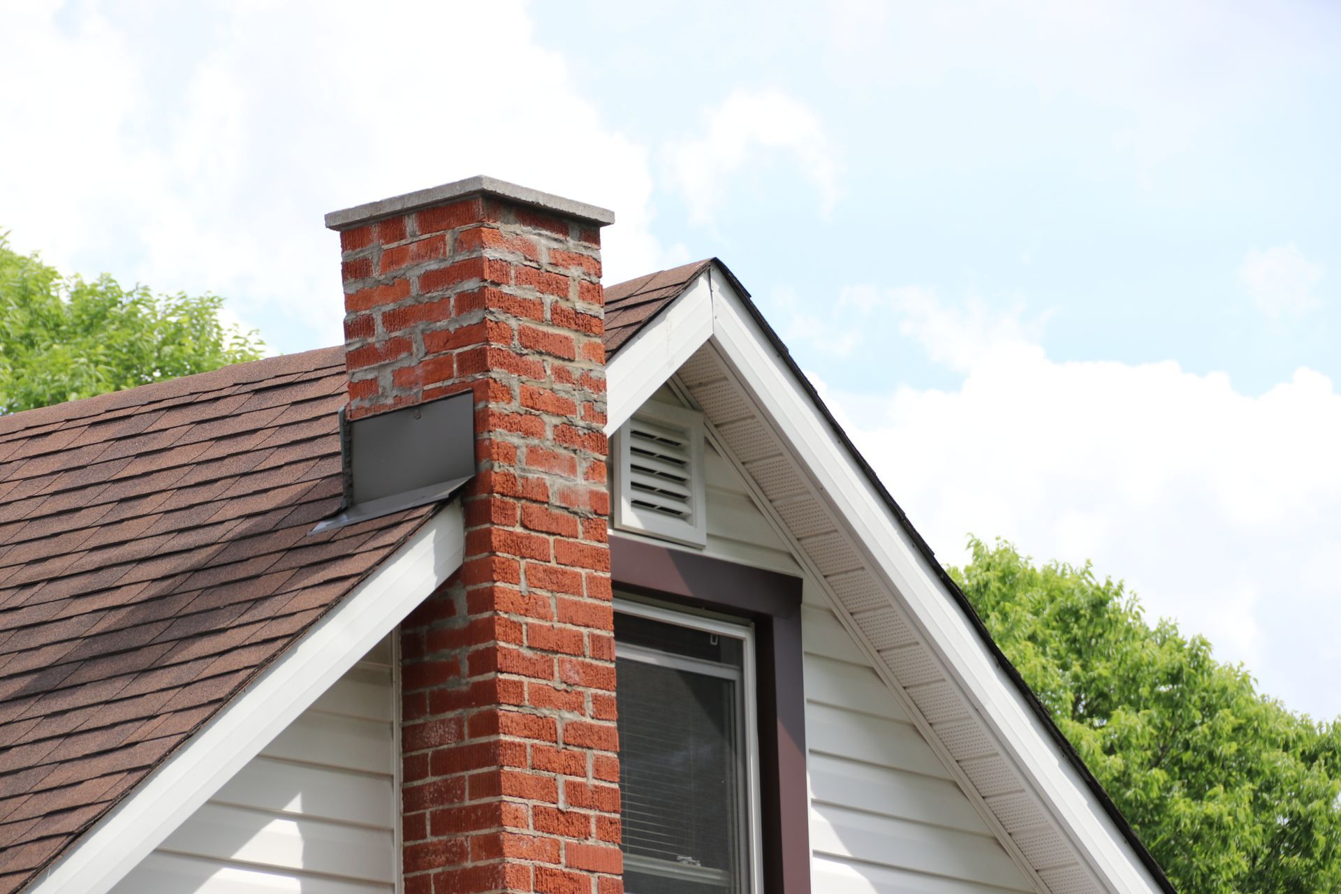 Brick chimney rising from a brown shingled roof, with white siding and a window.