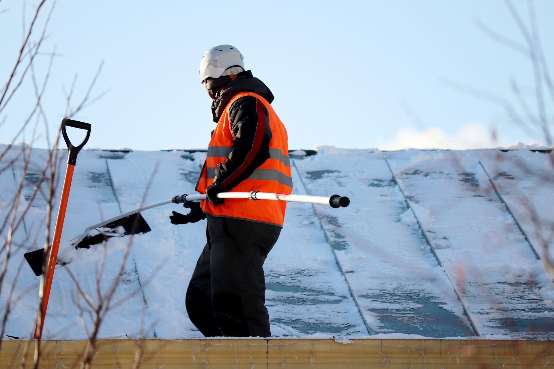 Person in safety vest and hard hat shoveling snow from a rooftop on a sunny day.