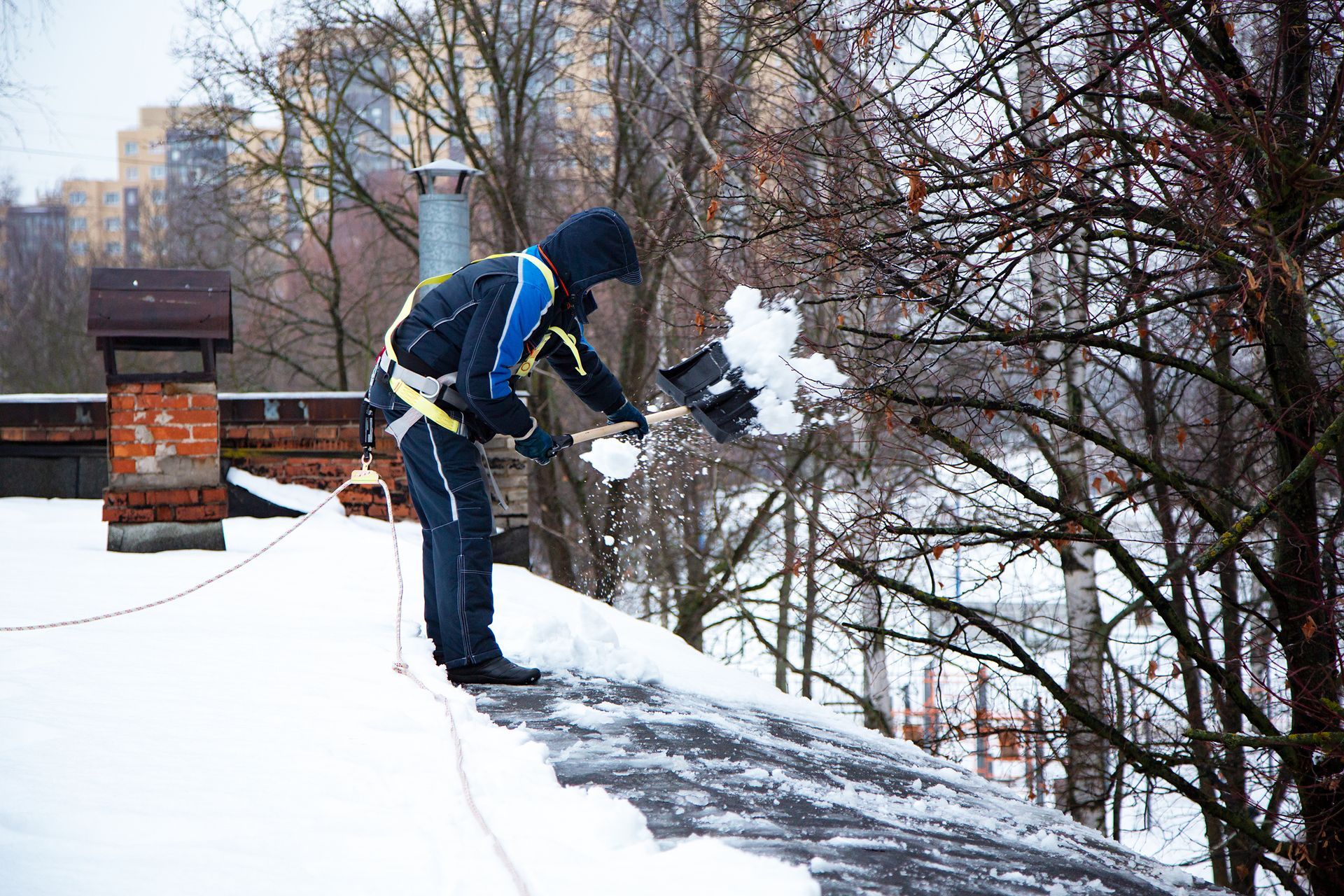 Une personne pelletant la neige d'un toit, portant un harnais de sécurité, sur fond urbain enneigé.