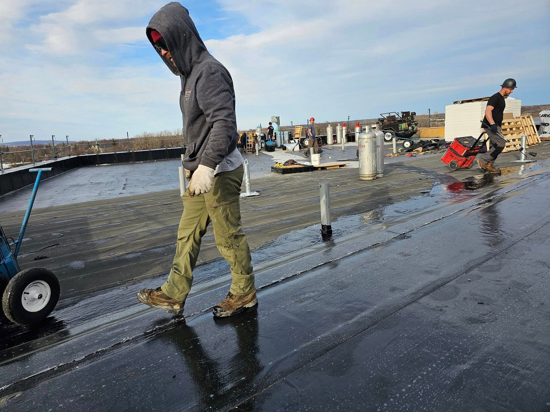 Un homme en sweat à capuche et pantalon de travail sur un toit plat, un autre utilisant une machine, journée ensoleillée.