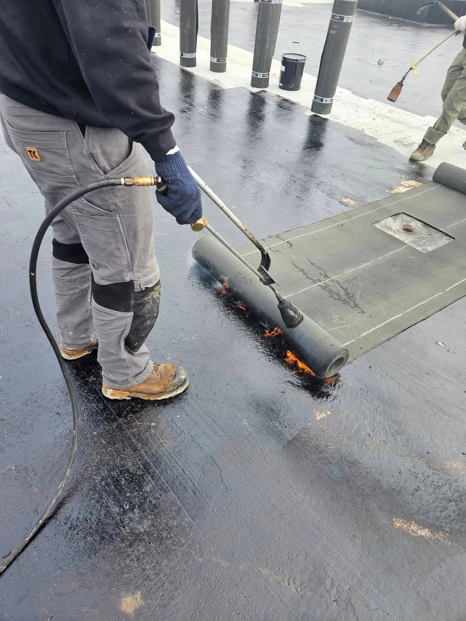 Personne utilisant une torche pour chauffer et sceller un matériau de toiture sur une surface sombre.